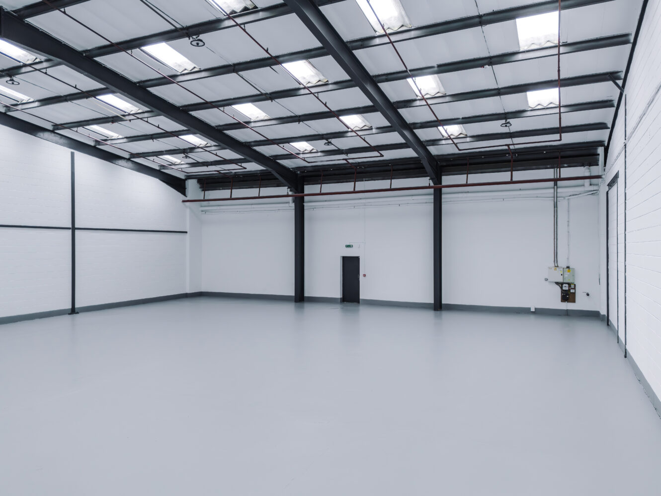 Empty industrial warehouse with white walls, high ceiling, metal beams, and a single closed door; the floor is smooth and grey. Bright overhead lights illuminate the space.