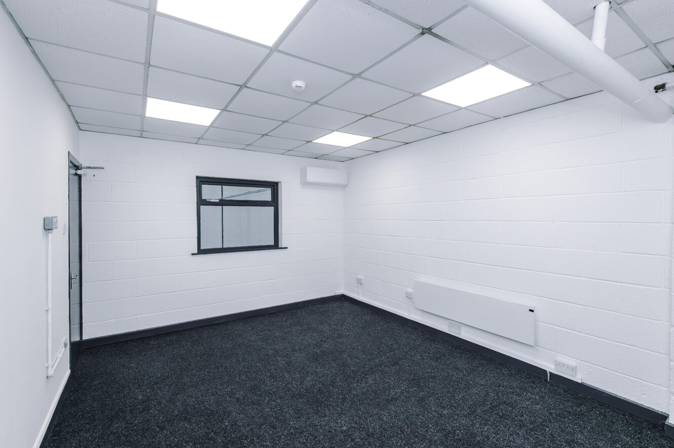 Empty office with white walls, a black-framed window, ceiling lights, dark carpeting, a closed door, and visible heating and air conditioning units.