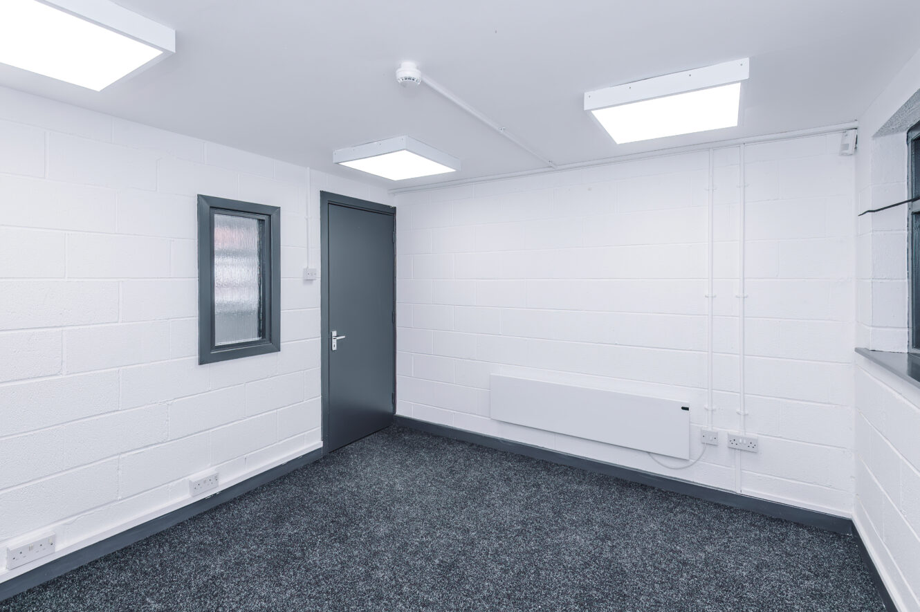Small empty room with white brick walls, a grey door, a window, wall heater, carpeted floor, and bright ceiling lights.