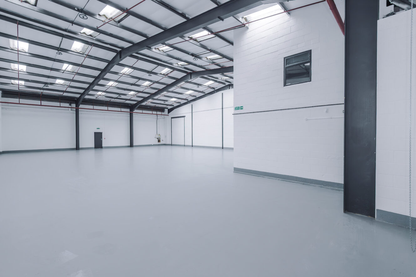 Interior of a large, empty industrial warehouse with white walls, grey floor, high ceiling, overhead lights, and exposed beams. A small room with a window is on the right side.