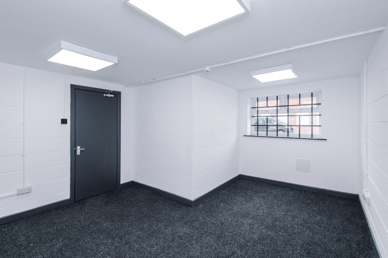 Empty white room with grey carpet, barred window showing a car outside, grey door, fluorescent ceiling lights, and white painted brick walls.