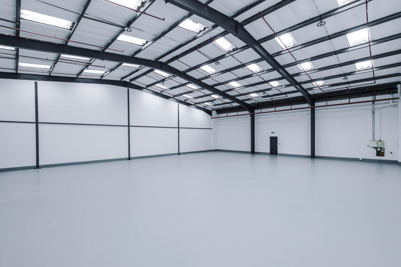 Spacious, empty industrial warehouse with white walls, high ceiling, exposed beams, skylights, and a single black door.