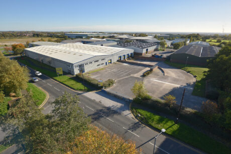 Aerial view of an industrial estate featuring large warehouses, parking areas, surrounding trees, and roads under a clear blue sky.
