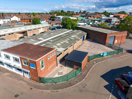 Aerial view of an industrial building complex with brick and metal structures, a gated entry, and an adjacent car park in a suburban area.