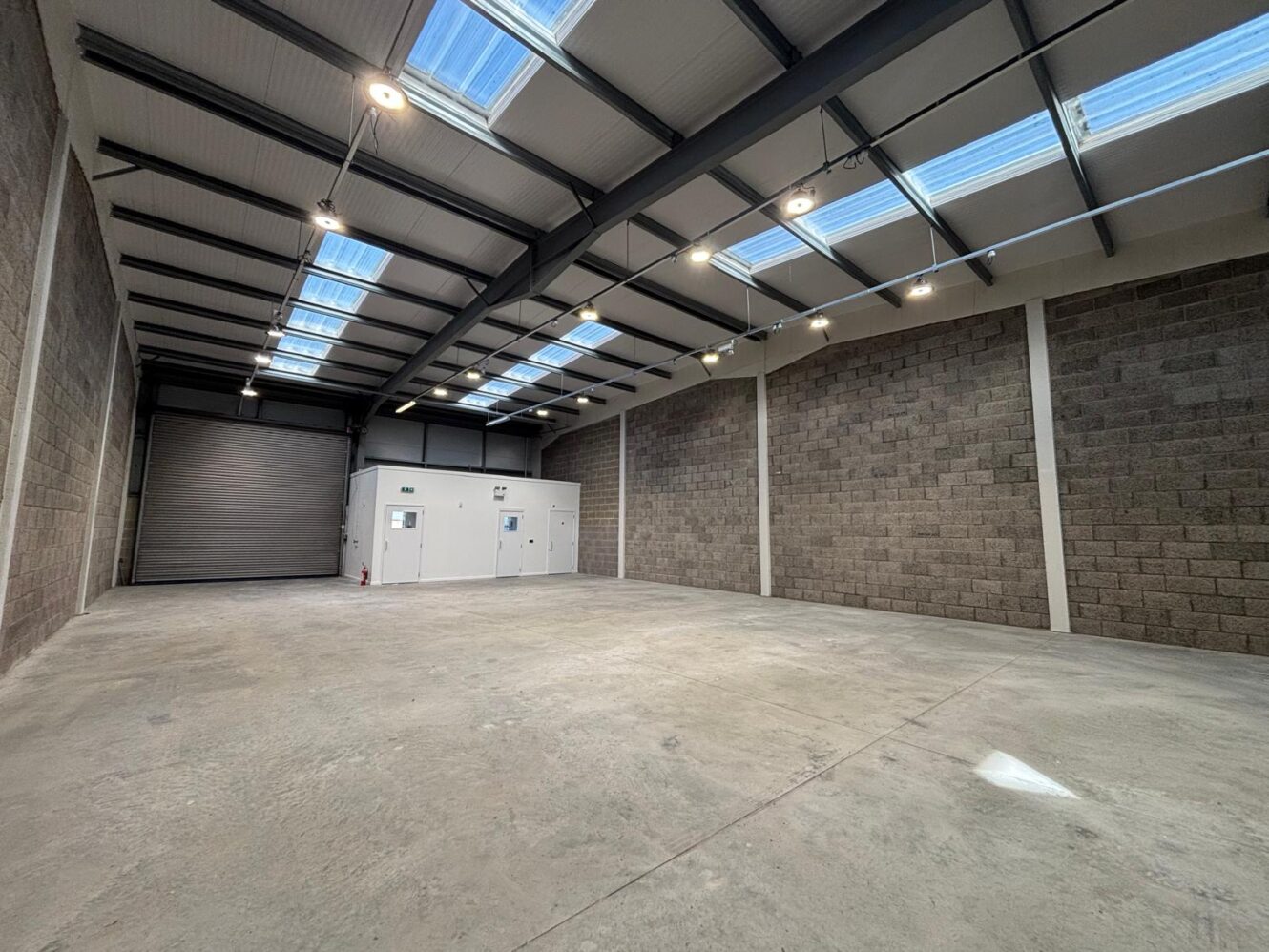 Empty industrial warehouse unit with a concrete floor, grey brick walls, and a large metal roller shutter door.