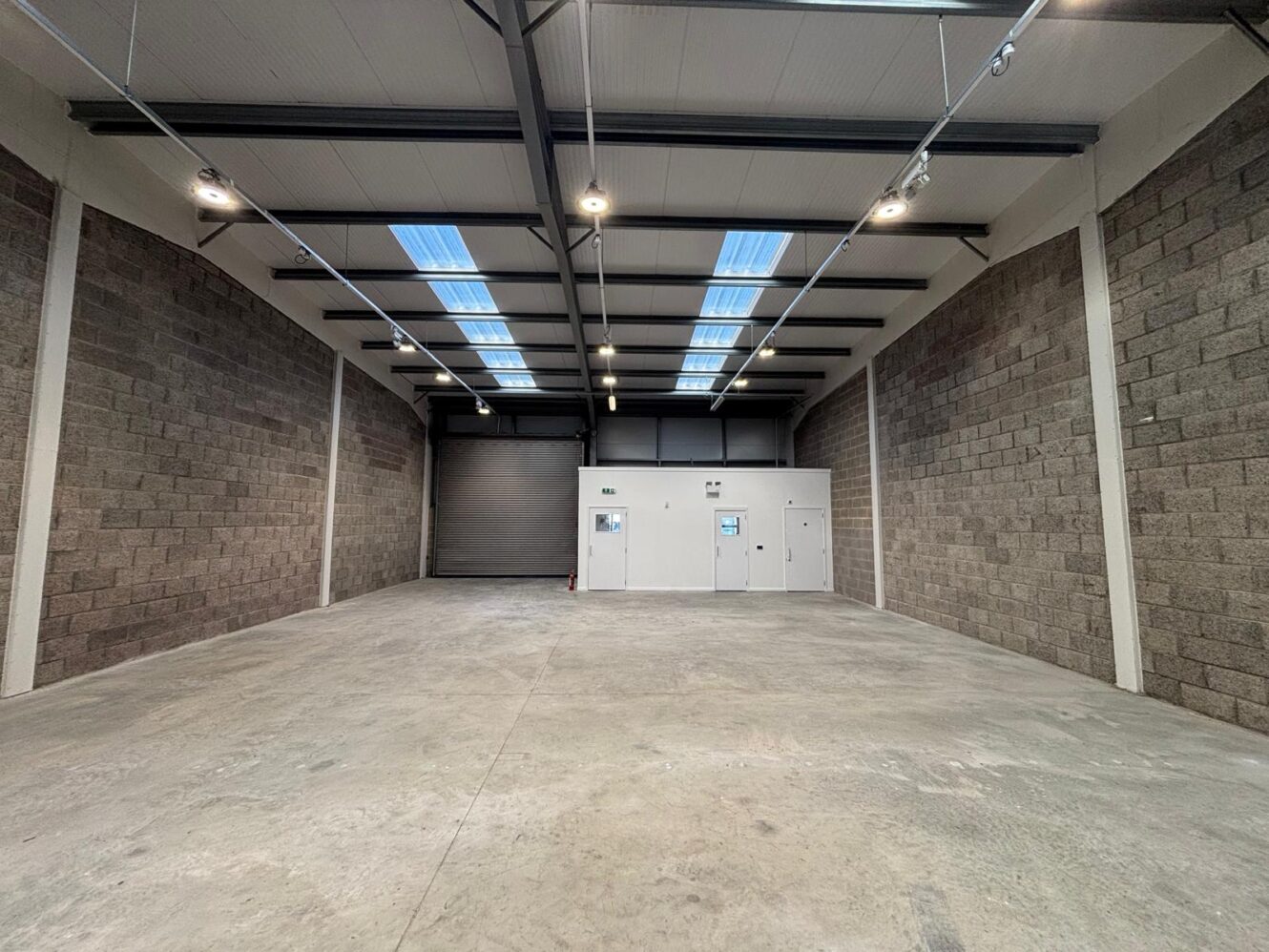 Empty warehouse space with grey concrete block walls, a concrete floor, a metal roller shutter door, and an interior office area.
