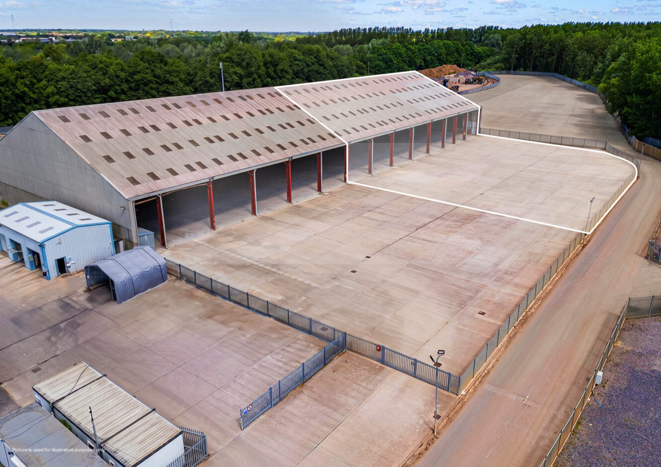 Aerial view of a large warehouse with open bays, an expansive concrete yard, and perimeter fencing.