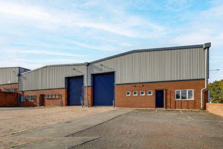 Industrial building with brick lower walls, grey metal upper walls, two blue roller shutter doors, and an adjacent car park.
