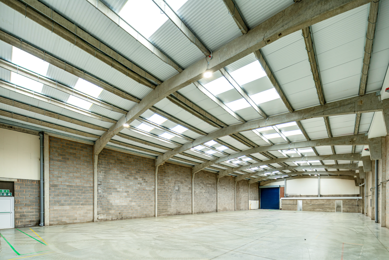 Large, empty industrial warehouse with concrete walls, a polished floor, and a metal roof featuring rows of skylights.