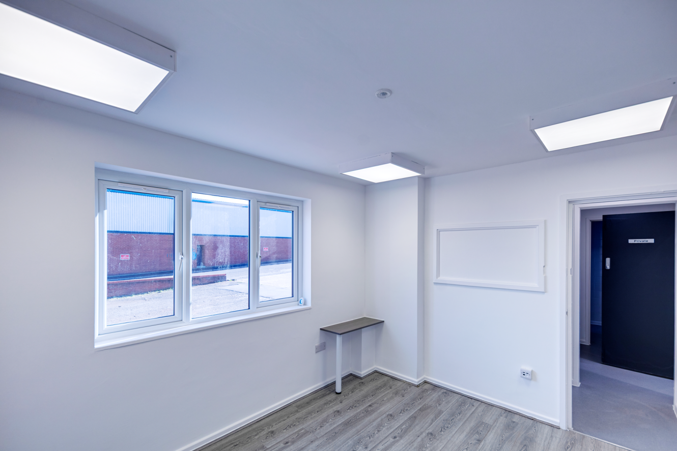 Empty office with white walls, a large window, a small wall-mounted desk, and a doorway leading to a room with a sign reading Private.