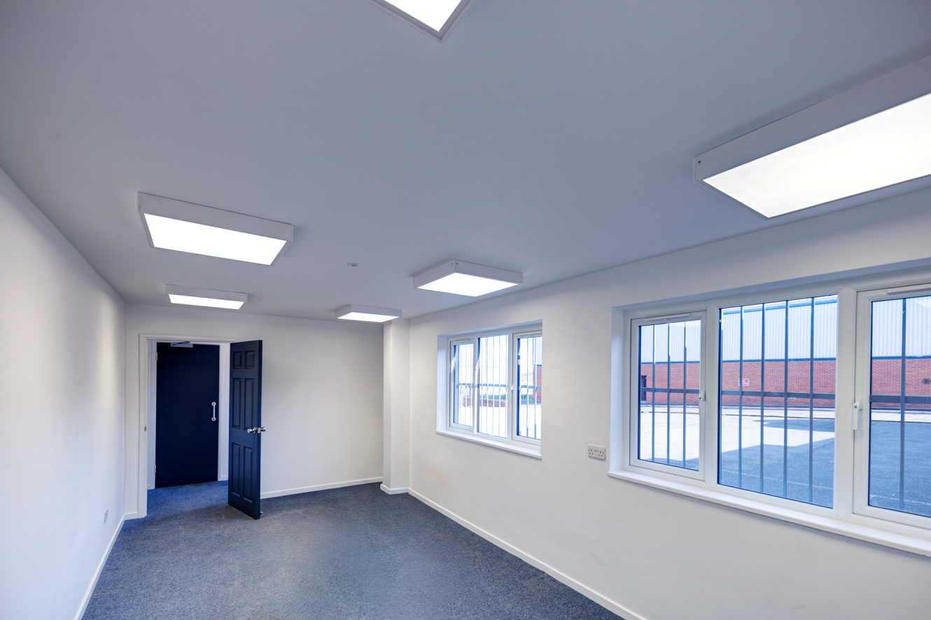 An empty room with white walls, grey carpet, several square ceiling lights, and windows with security bars looking out onto a car park.