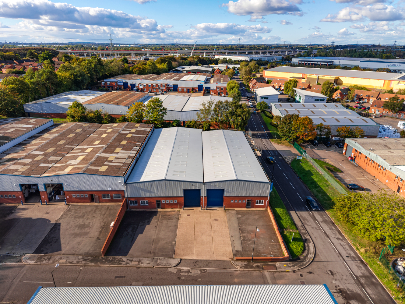 Aerial view of industrial warehouses and office buildings surrounding a road, with a large motorway flyover in the distance.