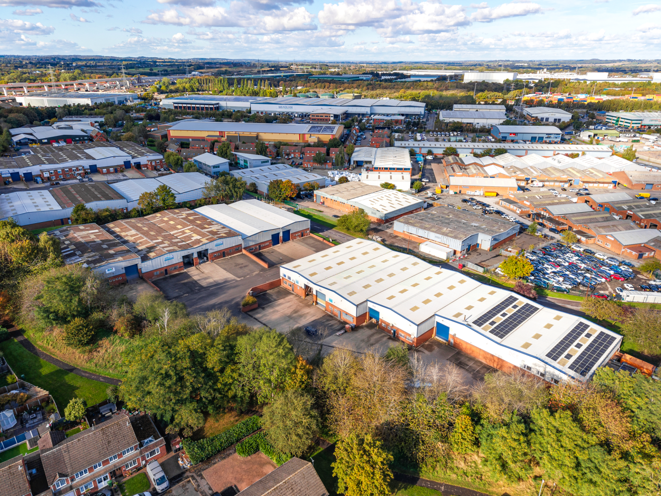 Aerial view of a sprawling industrial estate with multiple warehouse buildings, car parks filled with vehicles, and surrounding trees.