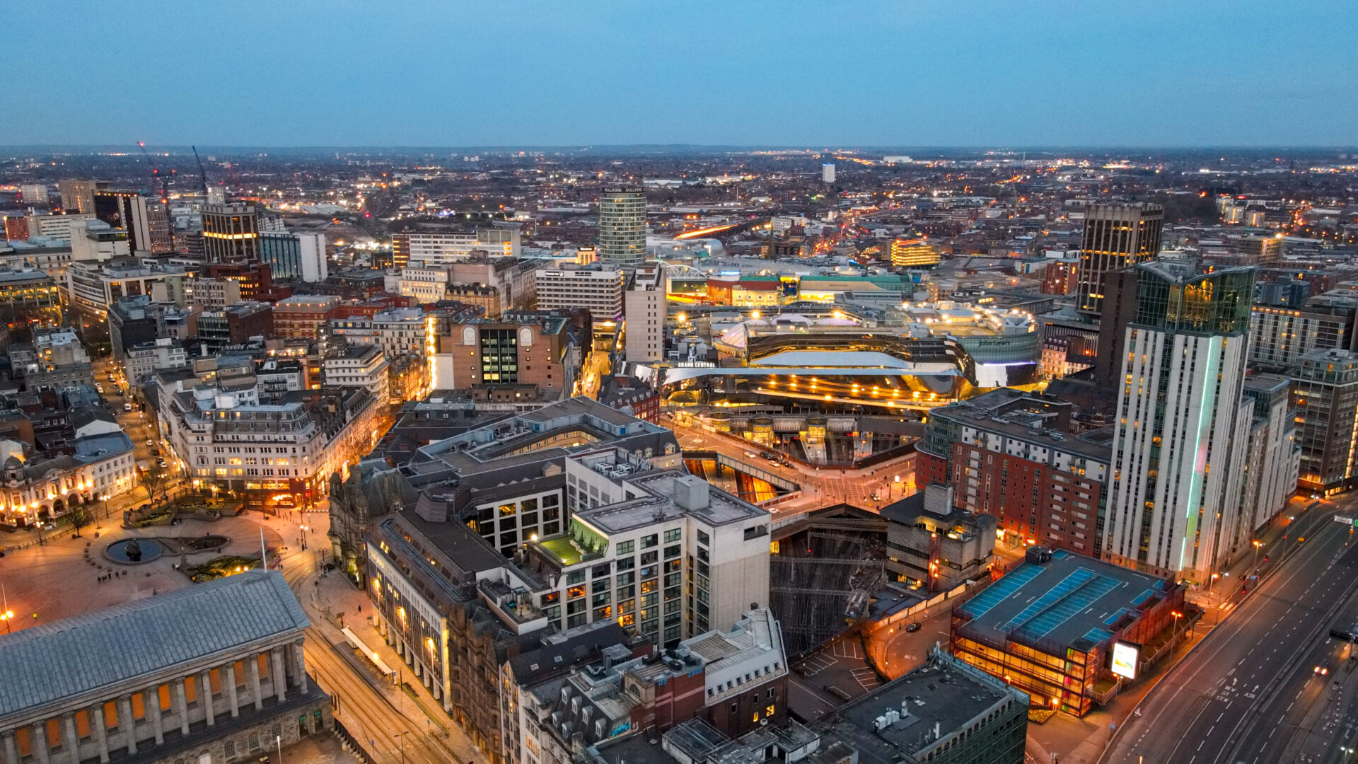 Aerial view of a city at dusk, showing illuminated buildings, streets, and the Grand Central shopping centre.