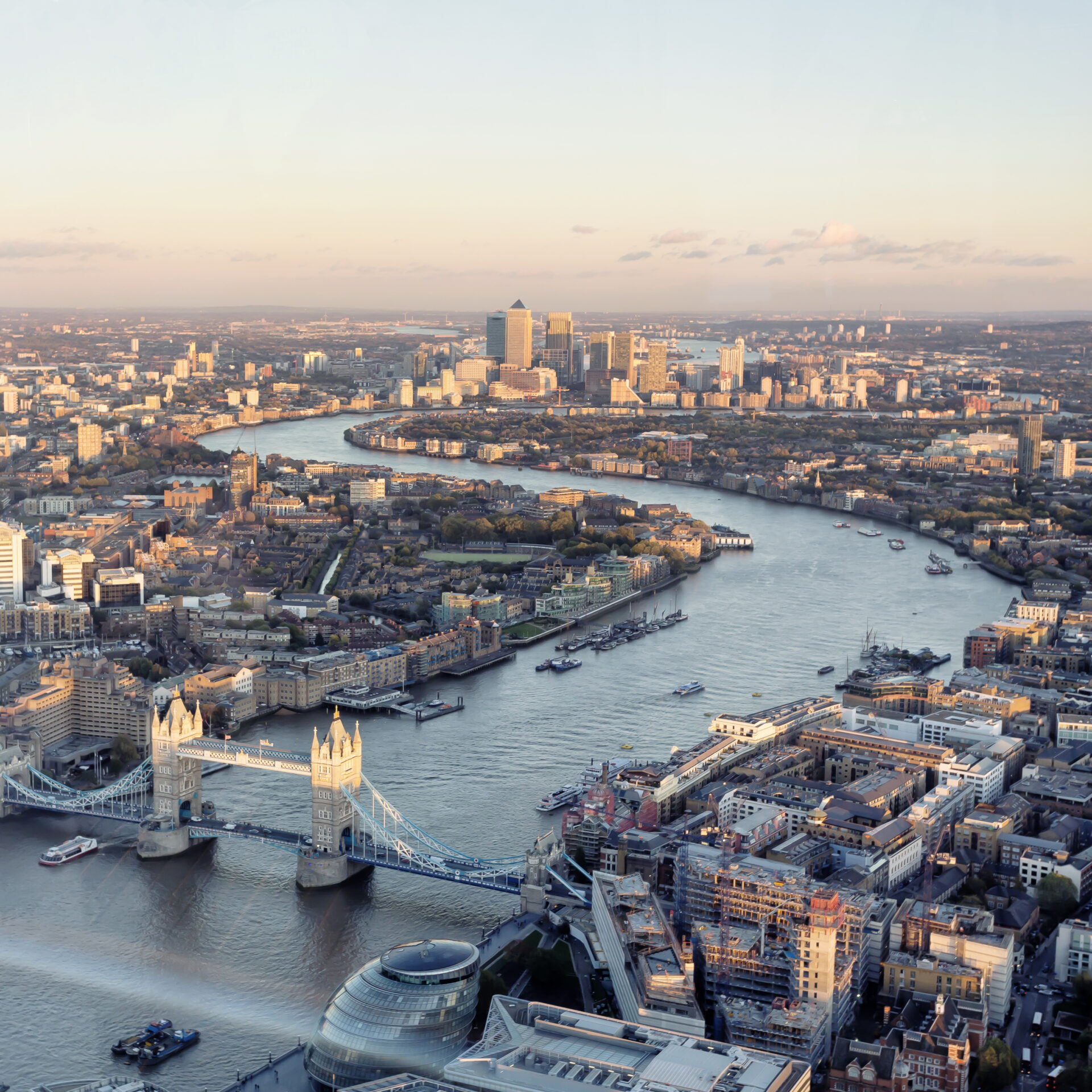 Aerial view of the River Thames winding through London, featuring Tower Bridge in the foreground and Canary Wharf in the distance.
