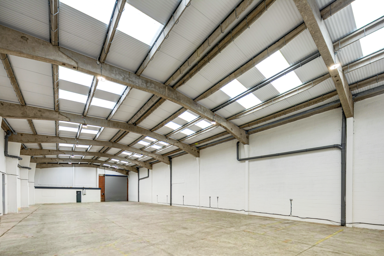 An empty industrial warehouse with white walls, a concrete floor, and a vaulted ceiling featuring numerous rooflights.