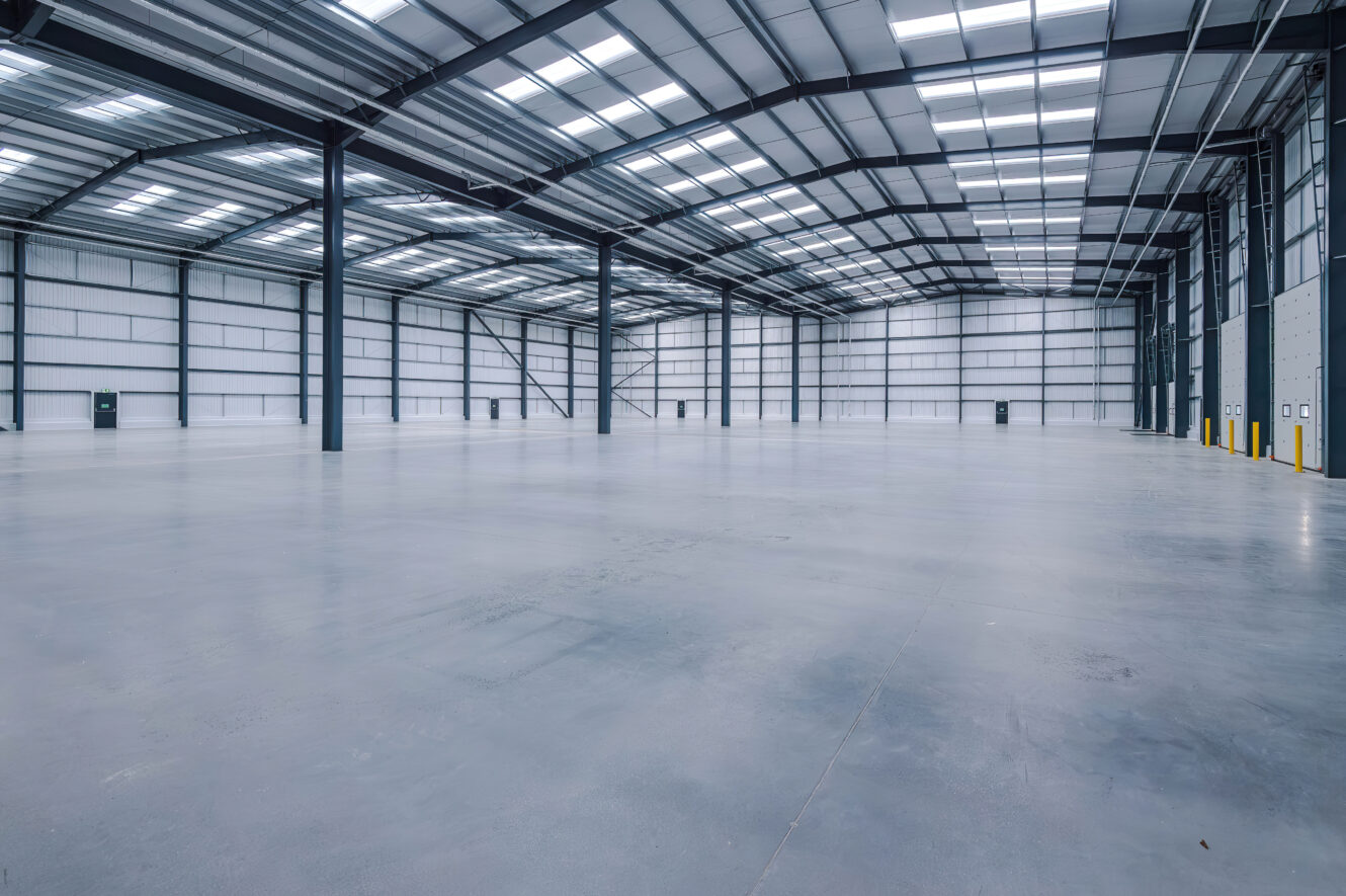 An expansive, empty industrial warehouse with a high steel-trussed ceiling, concrete floor, and evenly spaced support columns.