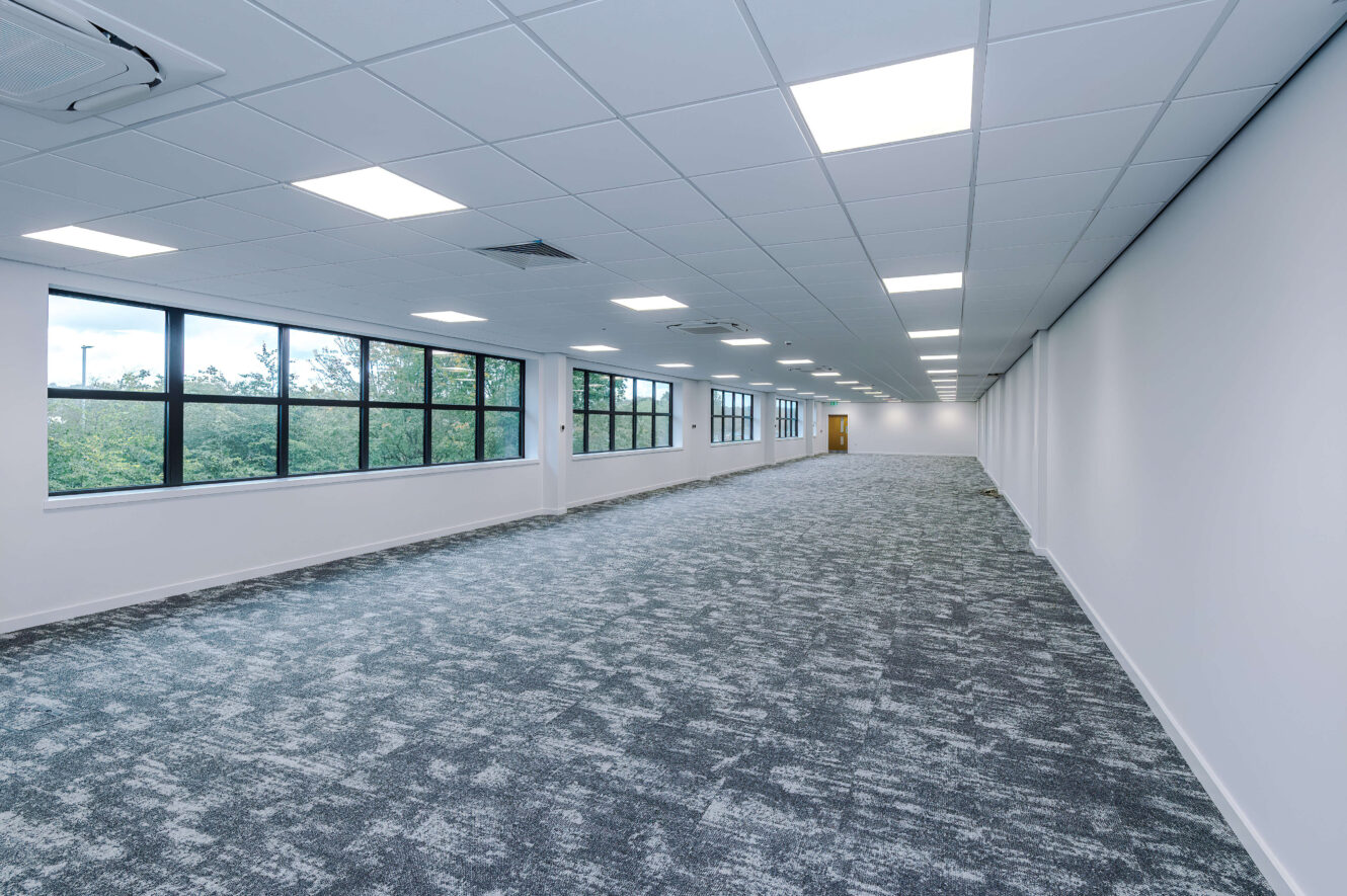 A large, empty office space with grey patterned carpeting, white walls, and a row of windows overlooking trees.