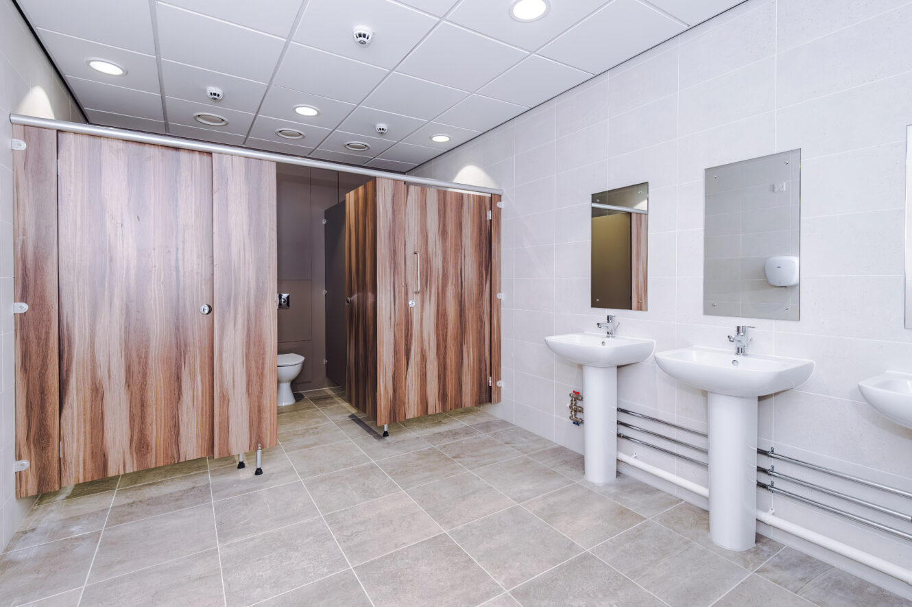 Public toilet with two wood-finish cubicles, two pedestal basins with mirrors, and tiled walls and floors.
