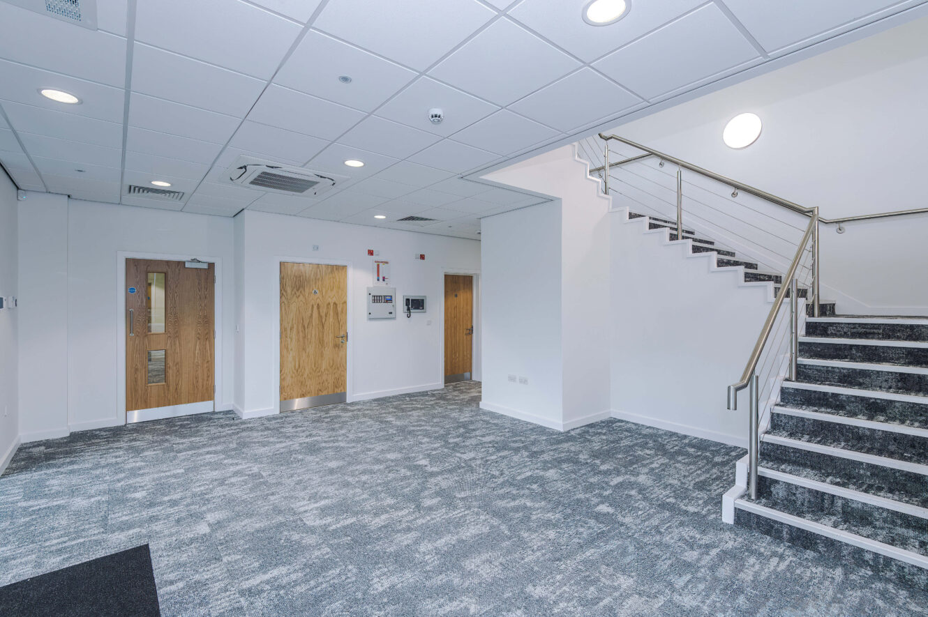 Modern office lobby with grey carpet, three wooden doors, and a staircase with a metal bannister.