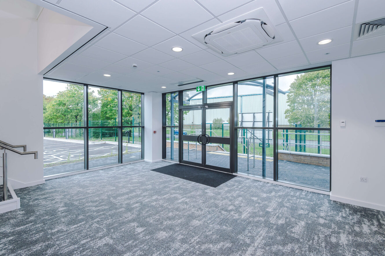 Empty office lobby with carpeted floors and floor-to-ceiling windows looking out onto a car park.