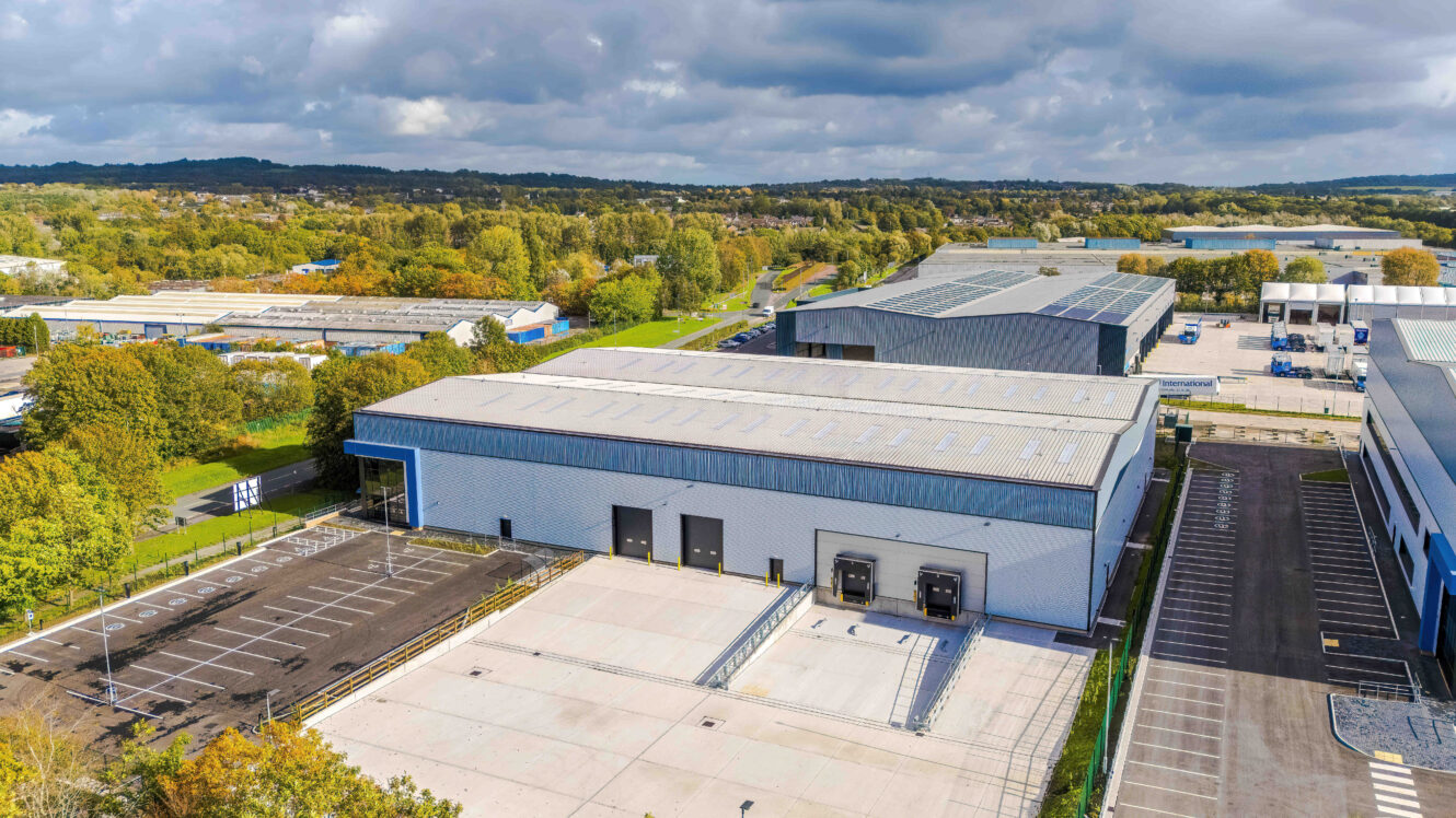 Aerial view of a large industrial warehouse with loading bays and a car park, surrounded by trees and other commercial buildings.