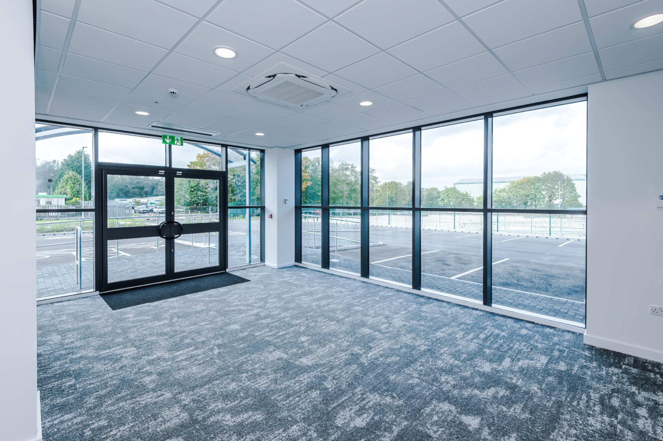 Empty office space with carpeted flooring, large glass windows, and a glass door leading to an outdoor car park.