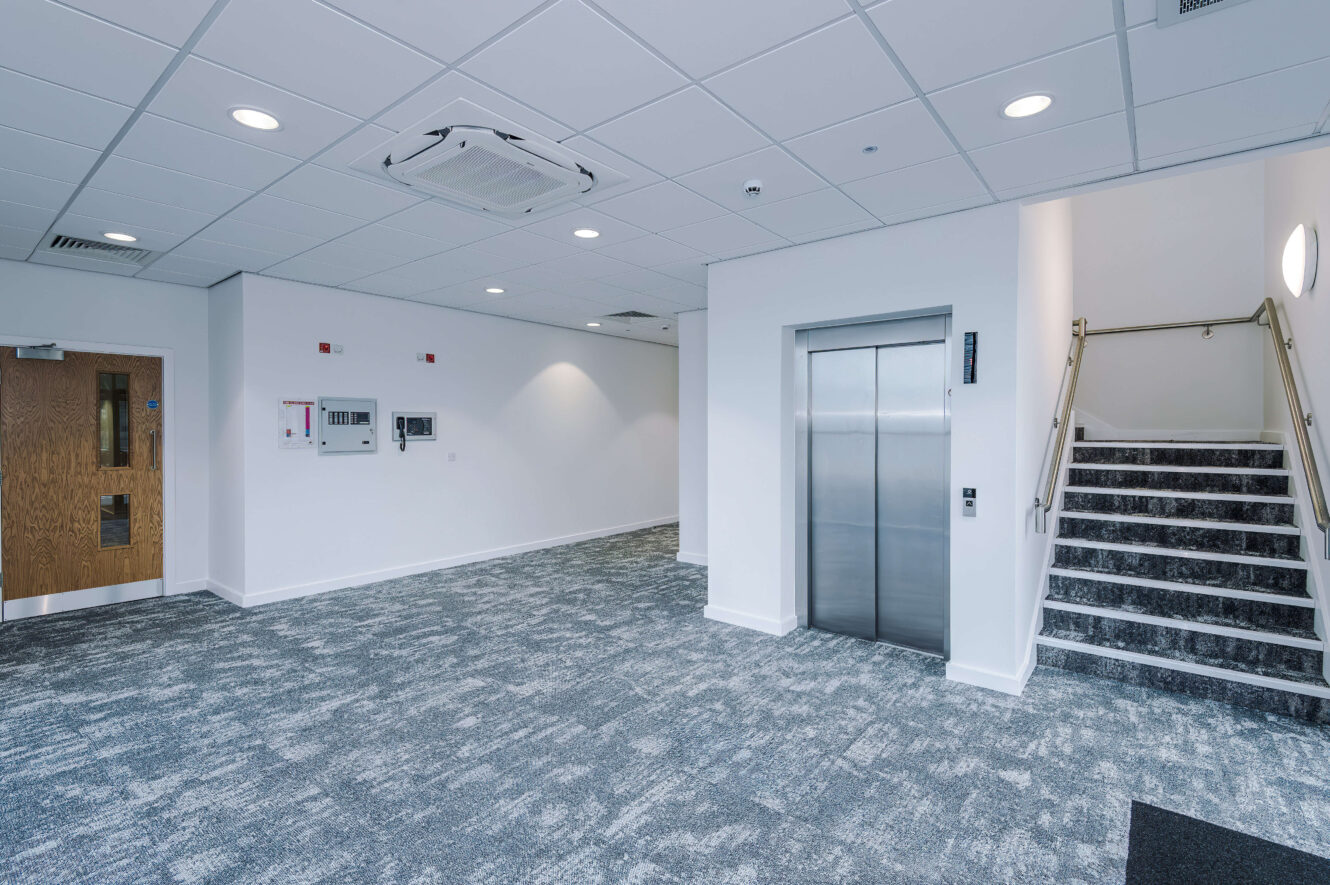 Office corridor with a wooden door, a metal lift, and a short staircase with a handrail.