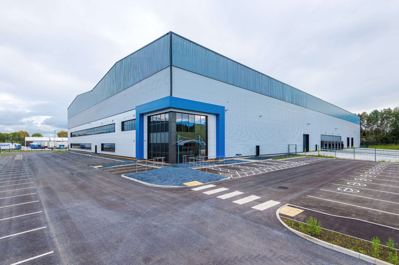 Large industrial warehouse building with metal cladding, blue trim, and a tarmacked car park in front.