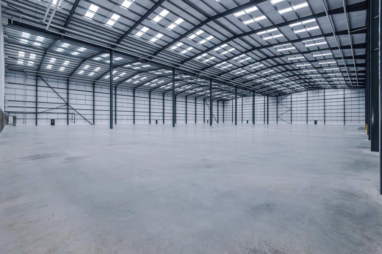 An empty, large-scale industrial warehouse with concrete floors, steel support beams, and rooflights.