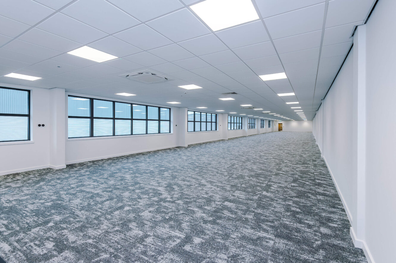 A long, empty office space with grey carpet and a grid ceiling featuring recessed lighting.