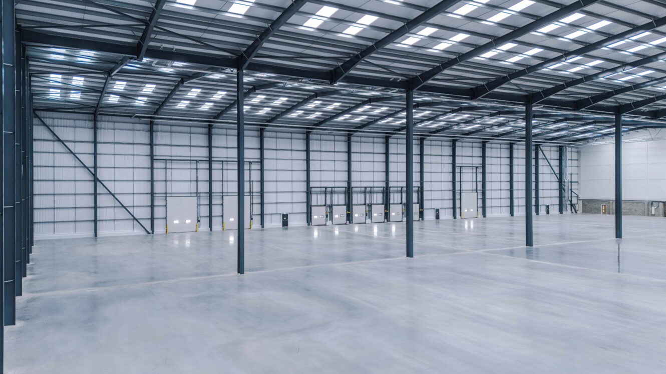 Interior of a large, empty warehouse with high ceilings, metal support beams, and several loading bay doors along the far wall.