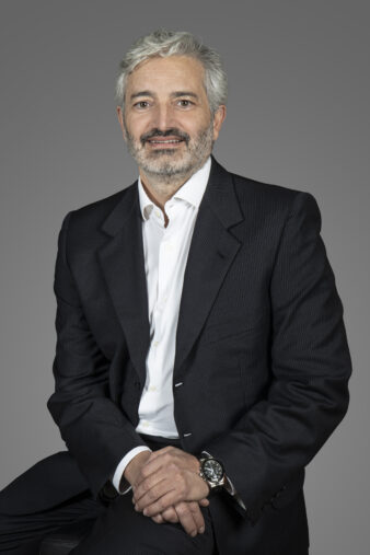 Un hombre de mediana edad, con pelo y barba canosos, vestido con traje oscuro y camisa blanca, sentado con las manos cruzadas sobre el regazo sobre un fondo gris liso.