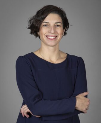 Una persona de pelo corto y oscuro, con un vestido azul marino, está de pie con los brazos cruzados y sonríe a la cámara sobre un fondo gris liso.