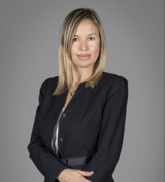 Una mujer de pelo rubio liso, vestida con una americana negra, está de pie sobre un fondo gris liso con los brazos cruzados.