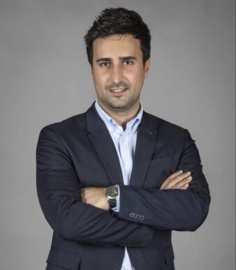 Hombre con traje oscuro y camisa clara de pie con los brazos cruzados, mirando a la cámara sobre un fondo gris liso.