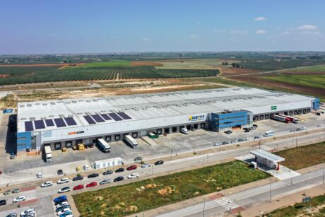 Una gran nave industrial con paneles solares en el tejado, múltiples muelles de carga y numerosos camiones y coches aparcados en el exterior, rodeada de campos.