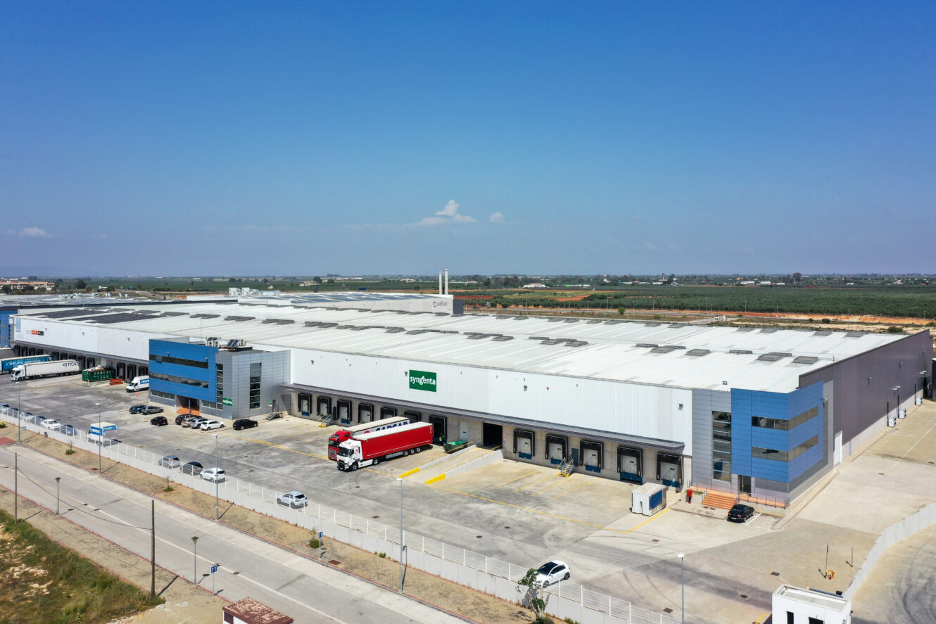 Una gran nave industrial con múltiples muelles de carga, camiones y coches aparcados delante, situada en una amplia zona abierta bajo un cielo azul.