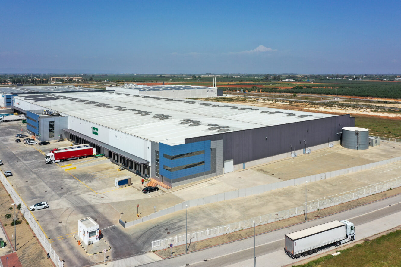 Una gran nave industrial con camiones aparcados en el exterior, rodeada de un terreno vallado y situada en una zona llana y rural bajo un cielo azul despejado.