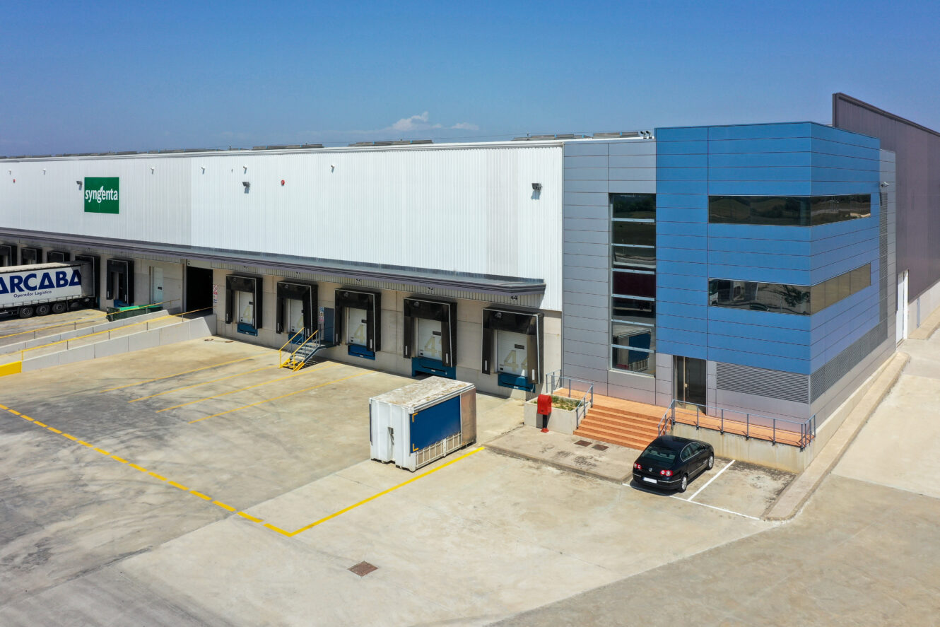 Una gran nave industrial con un exterior azul y blanco, múltiples muelles de carga, un coche negro aparcado y un camión blanco cerca de la entrada.