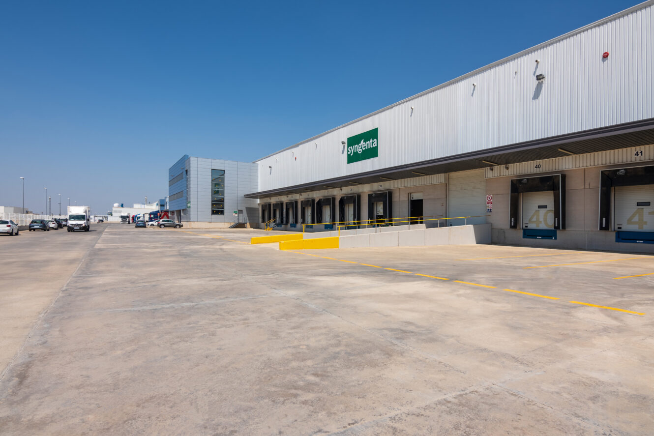 Una gran nave industrial con muelles de carga, rampa de acceso y un rótulo de Syngenta en el edificio, en un día despejado con cielo azul.