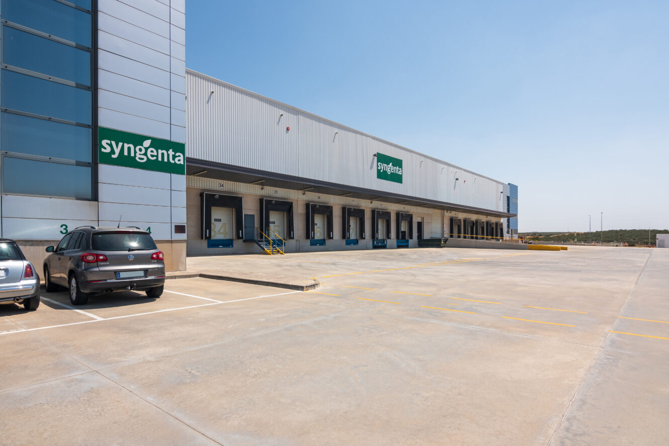 Un moderno edificio industrial con carteles de Syngenta, muelles de carga, coches aparcados y un aparcamiento vacío bajo un cielo despejado.
