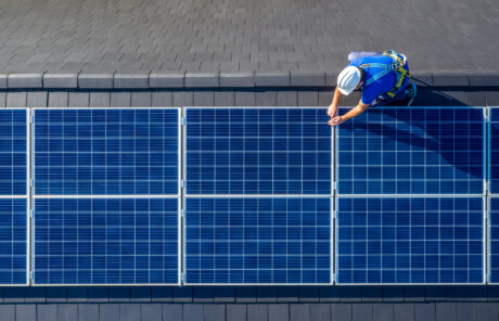 Un trabajador con equipo de seguridad instala o inspecciona paneles solares azules en un tejado inclinado gris oscuro, visto desde arriba.
