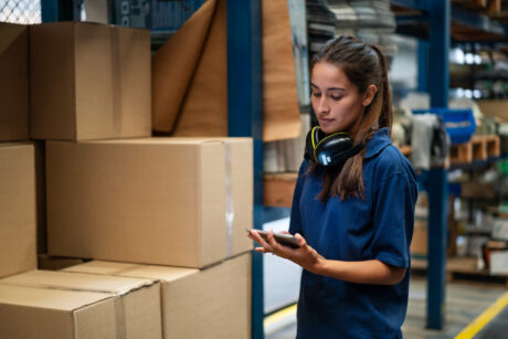 Una mujer en un almacén, de pie junto a cajas de cartón apiladas, sostiene una tableta digital con unos auriculares colgados del cuello.
