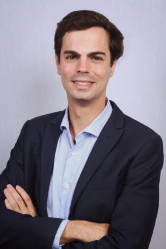 Un hombre con traje oscuro y camisa azul claro está de pie con los brazos cruzados, sonriendo delante de un fondo gris claro liso.