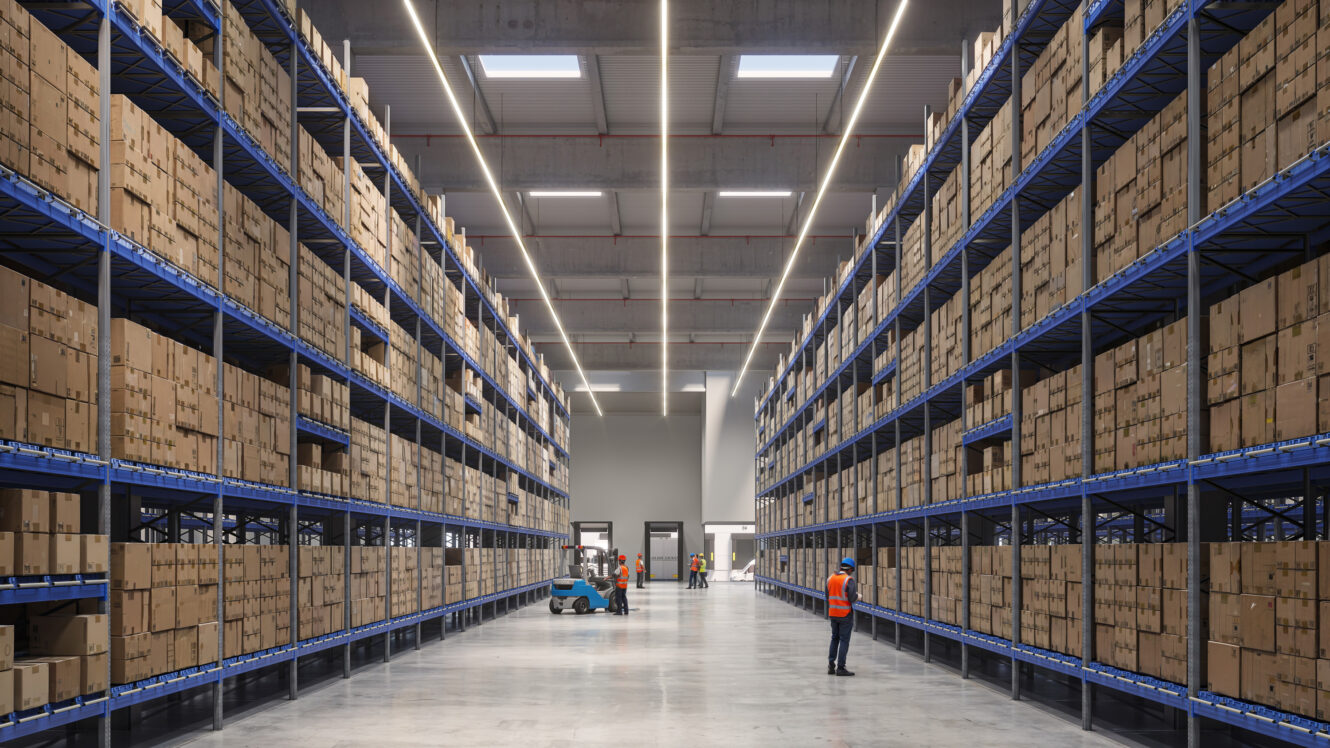 Interior de un gran almacén con estanterías altas apiladas con cajas de cartón, trabajadores con chalecos de seguridad y una carretilla elevadora operando en el pasillo central.