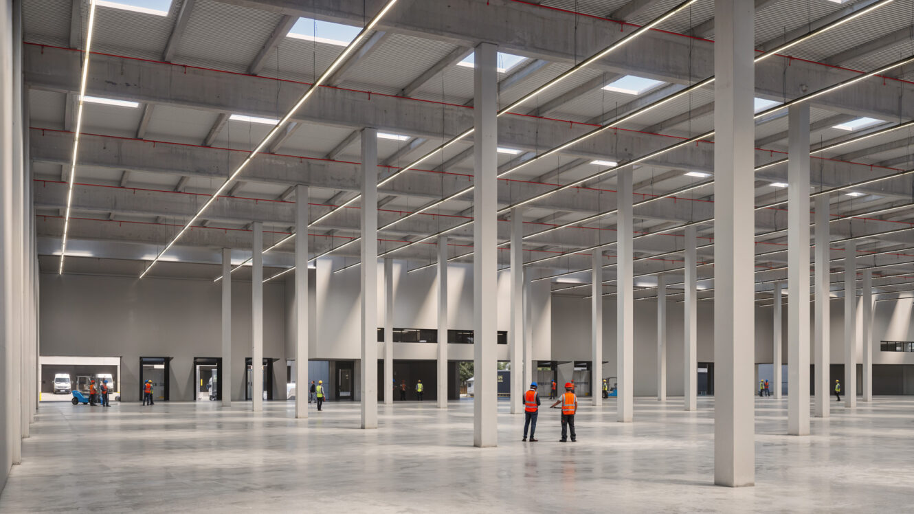 Un gran almacén vacío con techos altos, claraboyas y suelos de hormigón; varias personas con chalecos de seguridad están de pie y caminan por el interior.