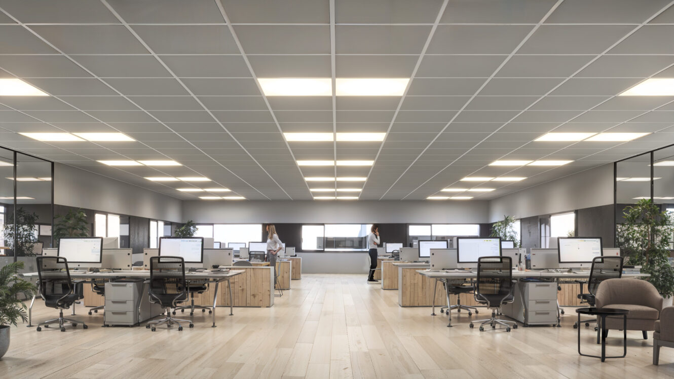 Moderna oficina diáfana con filas de mesas, ordenadores, sillas de oficina, plantas en macetas y dos personas de pie y caminando al fondo.