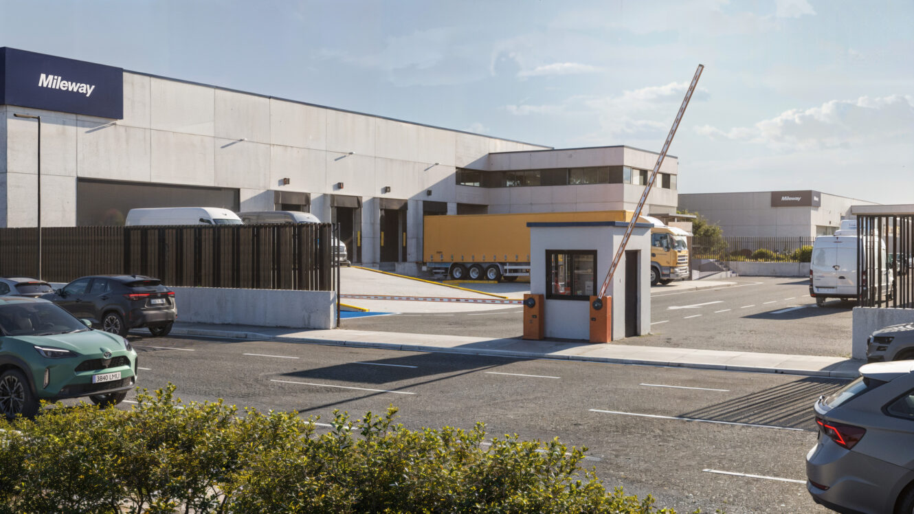 Un moderno complejo de almacenes industriales con camiones, coches, una puerta de seguridad y un brazo de barrera elevado, bajo un cielo azul despejado.