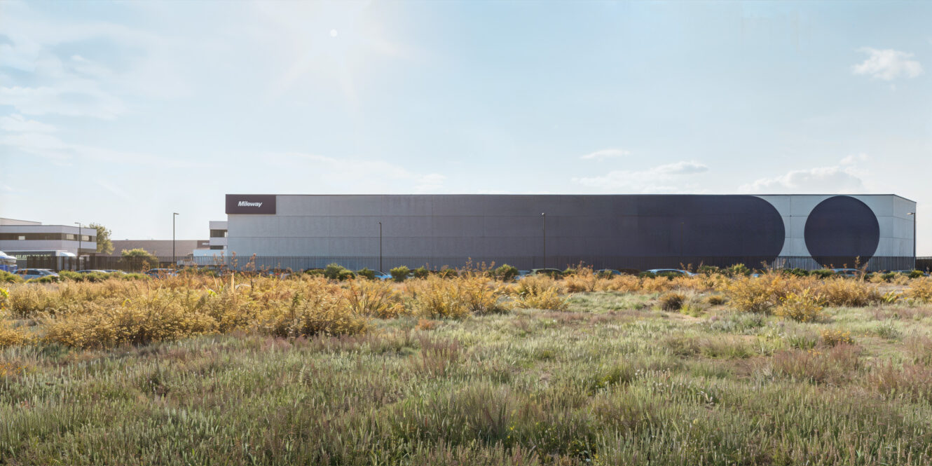 Detrás de un campo cubierto de hierba y arbustos dispersos, bajo un cielo casi despejado, se alza un gran edificio industrial moderno con un letrero en el que se lee Miltenyi.